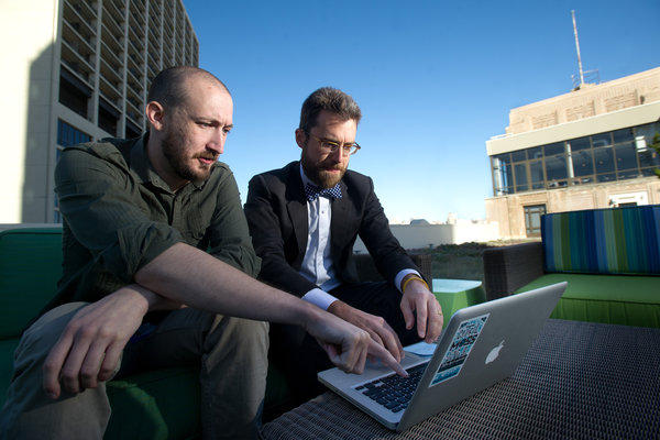 Two engineers on a roof deck in front of a keyboard. One bites his lip in concentration with hands on the keyboard while the other points at the laptop's screen